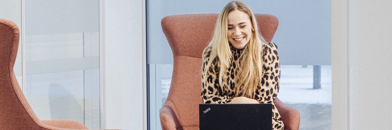 A young employee sits on an armchair in a modern, bright lounge area and works on a laptop on a small table in front of her.